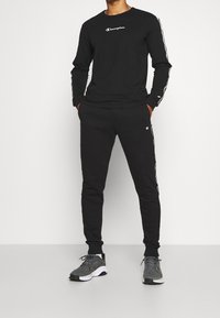 Schwarzes Langarmshirt und Sweatpants aus Baumwolle; mit Logo auf der Brust und vertikalen Streifen an den Ärmeln; kombiniert mit grauen Sportschuhen.