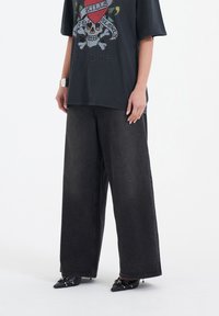Schwarzes Grafik-T-Shirt mit Totenkopfdesign, kombiniert mit weit geschnittenen schwarzen Denim-Hosen. Das Outfit wird durch spitze schwarze Schuhe ergänzt.
