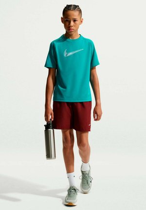 Junge Person mit geflochtenem Haar, die ein türkisfarbenes Nike-Shirt und rote Shorts trägt, hält eine silberne Wasserflasche und geht vorwärts auf weißem Hintergrund.