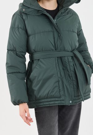 Personne portant une doudoune vert foncé avec une fermeture éclair à l'avant, des boutons pression, des poches latérales et une ceinture assortie nouée à la taille.