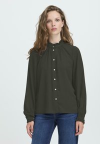 Femme avec des cheveux châtain clair ondulés portant un blouse noire à boutons avec un col et des poignets à volants, associée à un jean bleu, se tenant contre un fond pâle.