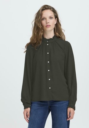 Eine Frau mit welligem, hellbraunem Haar trägt eine schwarze Bluse mit Knöpfen, die einen Rüschensaum und Rüschenmanschetten hat, kombiniert mit blauen Jeans und steht vor einem hellen Hintergrund.