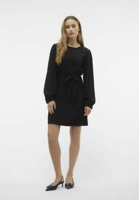 Črna obleka z teksturiranim pikčastim vzorcem, dolge naborkane rokave, okroglo ovratnico in pasom. Uporabljena s črnimi slingback petami.