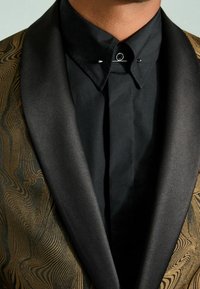 Chaqueta de satén negro con cuello de chal y solapa texturizada con patrón en dorado y marrón, combinada con una camisa negra de botones con un alfiler.