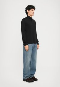 Mann med krøllete mørkt hår som har på seg en svart langermet skjorte, løse blå jeans og mørkebrown sko, står mot en enkel hvit bakgrunn.