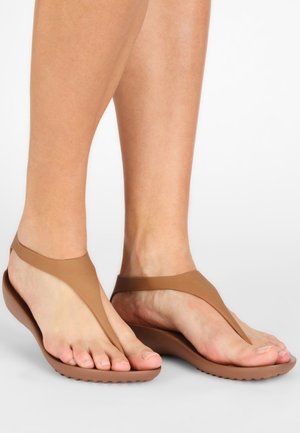 Fødder iført brune thongsandaler med en glat rem og let formet sål på en hvid baggrund.