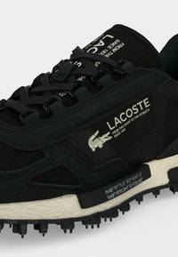 Μαύρο αθλητικό παπούτσι Lacoste με λευκή σόλα που έχει το λογότυπο της μάρκας, κείμενο και γεωγραφικές συντεταγμένες στο πλευρό και τη γλώσσα.