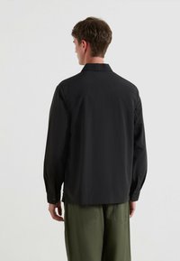 Mann in einem schwarzen Langarmshirt und olivgrünen Hosen, der mit dem Rücken zu einem schlichten hellen Hintergrund steht.