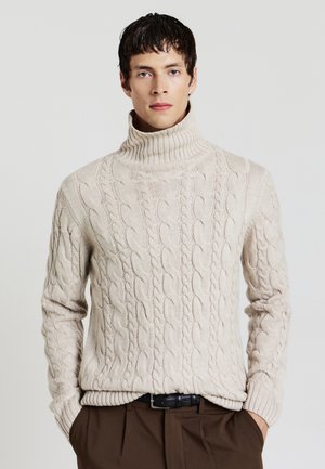 Pull à col roulé en tricot torsadé beige avec un ourlet et des poignets côtelés, présentant un motif texturé et une coupe décontractée. Associé à un pantalon marron.