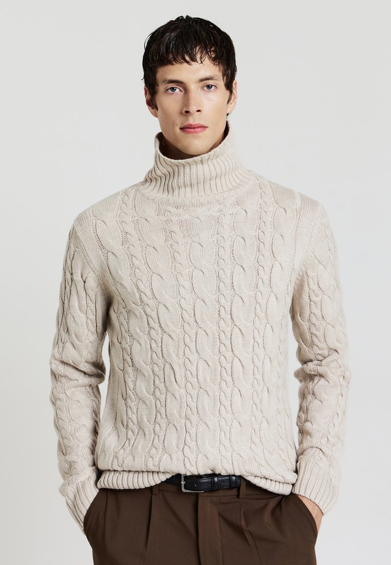 Beige Kabelstrick-Pullover mit Rollkragen, ribbelausschnitt und -ärmeln, einem strukturierten Muster und einer lockeren Passform. Kombiniert mit braunen Hosen.