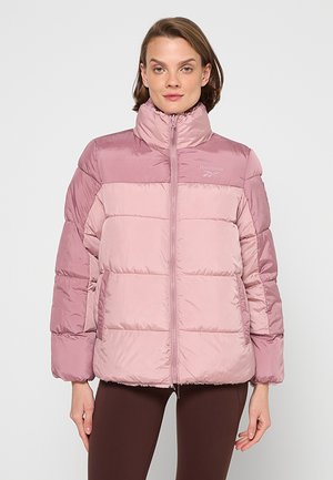 Donna che indossa una giacca imbottita Reebok rosa chiaro e polveroso, chiusa con cerniera, in piedi su uno sfondo bianco semplice.