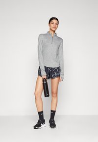 Graues Langarmoberteil mit einem Reißverschluss am Hals, gemusterte marineblaue Shorts, schwarze Wasserflasche, schwarze Turnschuhe und schwarze Socken mit weißen Akzenten.