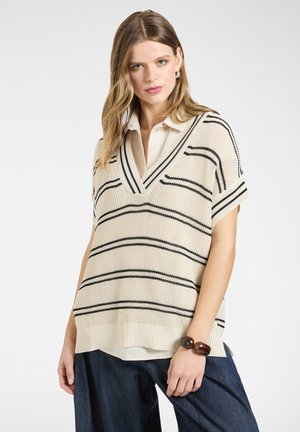 Jonge vrouw die een crèmekleurig gebreid vest met zwarte strepen draagt over een wit shirt en donkerblauwe broek, gecompleteerd met een bruine armband.