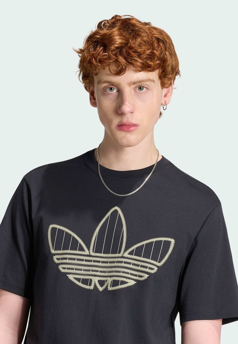 Jeune personne aux cheveux roux bouclés portant un T-shirt noir avec un grand logo Adidas beige brodé et un collier chaîne en argent.