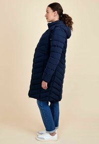 Geacă puffer bleumarin cu glugă, având un model în formă de zigzag și un fermoar frontal. Purtată peste blugi albaștri și adidași albi.