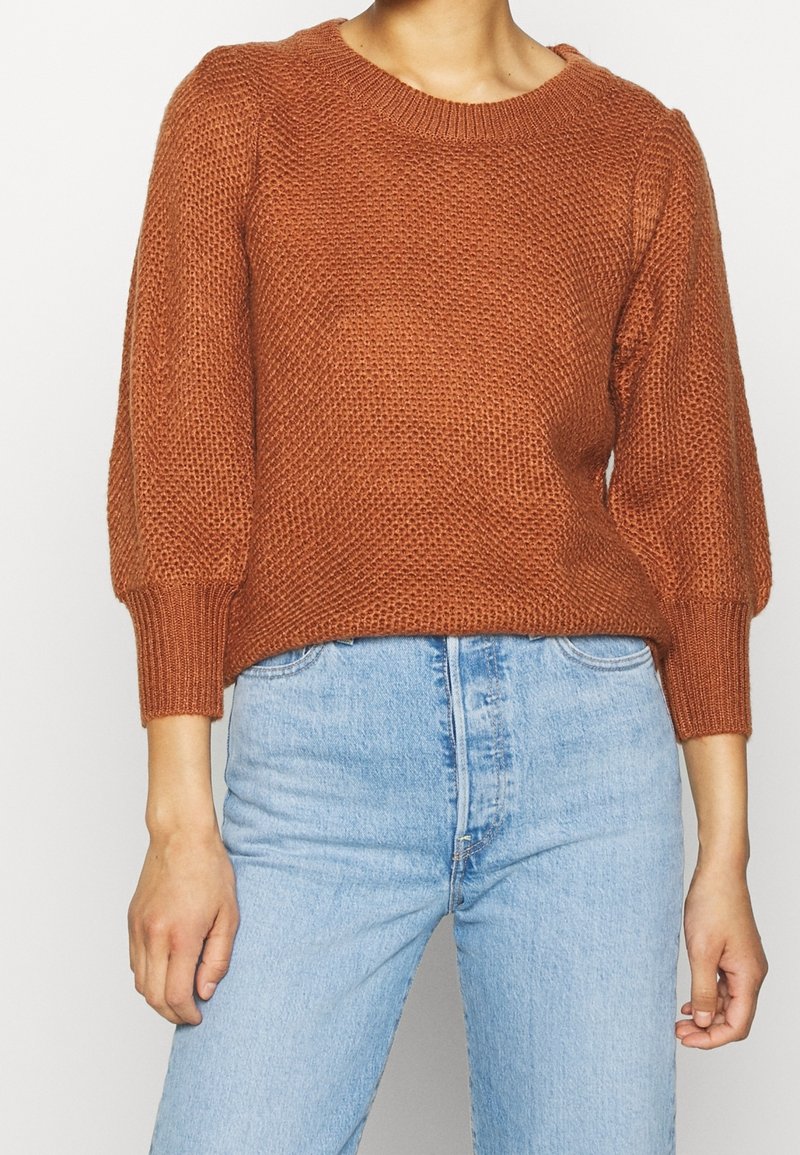 Getextureerde gebreide trui in roestoranje, met een ronde halslijn en driekwart pofmouwen, gecombineerd met lichtblauwe denim jeans.
