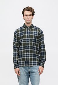 Fiatal férfi gombolt sötétzöld és tengerészkék kockás ingben, világoskék farmerral, egy sima fehér háttal áll.