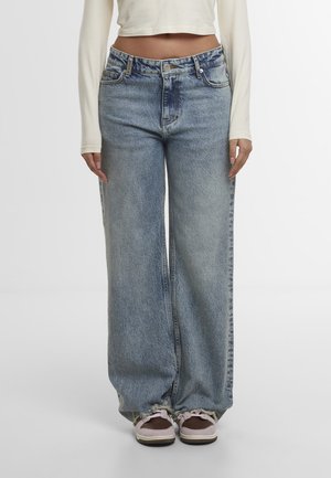 Person wearing high-waisted, wide-leg blue jeans with frayed hems and pink sneakers, paired with a cropped long-sleeve white top.