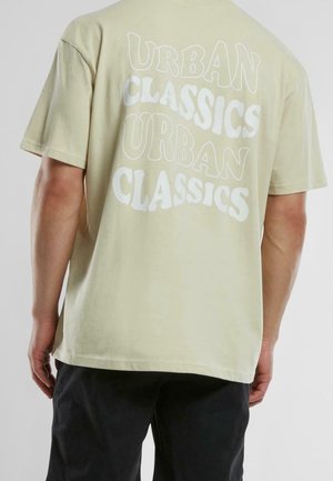 Person trägt ein hellbeiges T-Shirt mit dem Schriftzug "URBAN CLASSICS", der zweimal in Weiß auf dem Rücken wiederholt ist, kombiniert mit einer schwarzen Hose.