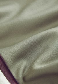 Textil i dämpad olivgrön, med en slät, texturerad yta med svag lyster. Kanterna har en kontrasterande mörkröd kant.