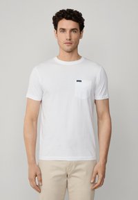 Camiseta de algodón blanca con cuello redondo, mangas cortas y un bolsillo en el pecho izquierdo con una etiqueta azul marino. Diseño simple y clásico.