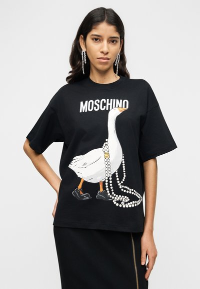Μαύρο βαμβακερό μπλουζάκι με γραφικό λευκής πάπιας που φοράει μαύρα παπούτσια και περιδέραιο με μαργαριτάρια. Στο μπροστινό μέρος περιλαμβάνει το κείμενο "MOSCHINO" πάνω από το σχέδιο.