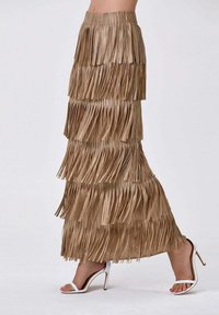 Jupe maxi à franges en beige, design superposé avec des détails horizontaux. Présente une surface texturée et une taille élastique. Portée avec des talons blancs.