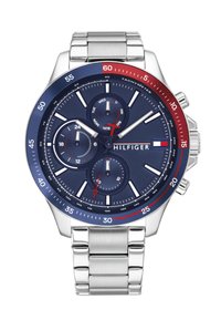 Blue dial Tommy Hilfiger wristwatch with silver metal band, chronograph subdials, red and blue bezel, and day, 24-hour, and seconds indicators.