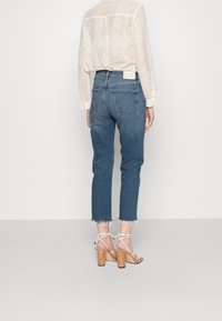 Vaqueros de mezclilla en un azul intermedio con un corte recto, dobladillo deshilachado y dos bolsillos en la parte trasera. Combinados con sandalias de tacón blancas con tiras.