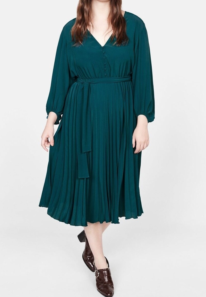Femme portant une robe midi plissée vert foncé à manches longues et boutons, associée à des chaussures à talons marron foncé.