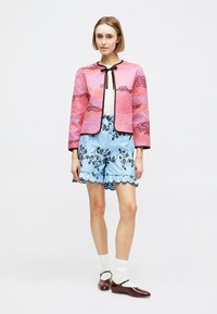 Γυναίκα που φοράει ροζ υφασμάτινο σακάκι με μαύρη επένδυση, μπλε σορτς με κεντήματα λουλουδιών και βολάν στη λαιμόκοψη, λευκές κάλτσες και σκούρα καφέ παπούτσια Mary Jane.