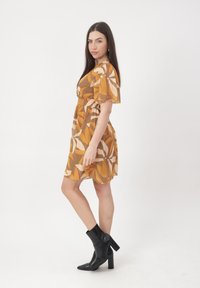 Mujer de pie de lado, vestida con un vestido corto de estampado floral en tonos naranja y beige, y botas negras de tacón hasta el tobillo, sobre un fondo blanco liso.