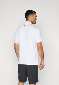 Hvid polo t-shirt med krave og korte ærmer, der har et sort logo på bagsiden af halsudskæringen. Kombineret med mørke shorts, hvilket giver et afslappet look.