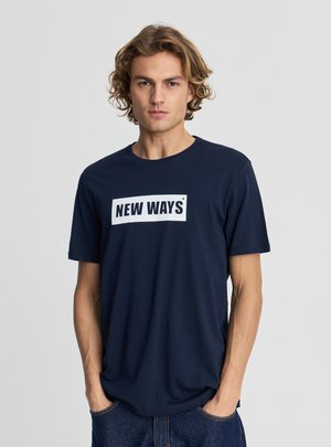 Młody mężczyzna o kręconych blond włosach, ubrany w granatowy T-shirt z napisem „NEW WAYS”, stoi z rękami w kieszeniach na jednolitym tle.