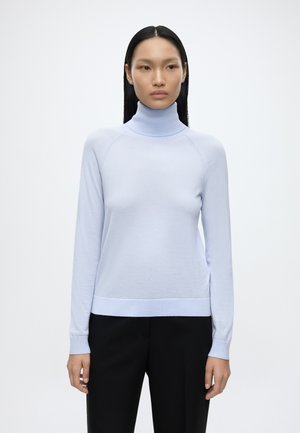 Kvinde med langt sort hår iført lyseblå højhalset sweater og sorte bukser står mod en ensfarvet baggrund.
