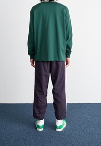 Person som står med ryggen mot kameran, klädd i en grön långärmad tröja, mörka byxor med vita sidostrippar, vita strumpor och gröna sneakers på en blå matta.