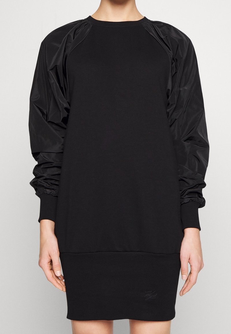 Femme portant une robe noire à manches longues avec des manches bouffantes brillantes et des poignets côtelés sur un fond clair uni.