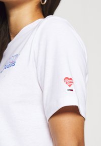 T-shirt en coton blanc avec un logo bleu sur le devant et un graphique en forme de cœur rouge "Tommy Jeans" sur la manche gauche.
