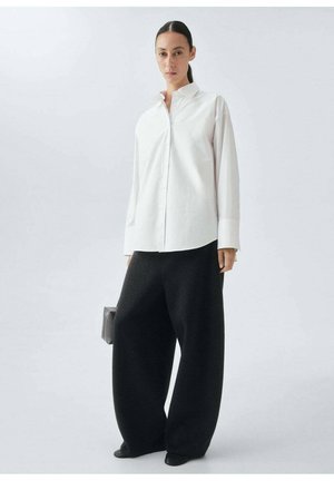 Camicia bianca in cotone con colletto, abbinata a pantaloni neri in lana a gamba larga. Il modello tiene una piccola borsa nera.