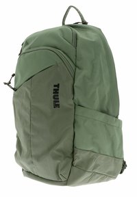 Mochila de tela verde con un acabado texturizado, que cuenta con un bolsillo lateral, detalle del logo y un asa de transporte reforzada en la parte superior.