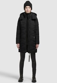 Zwarte pufferjas met een capuchon, mooi gewatteerd patroon en ritssluiting aan de voorkant. Gecombineerd met zwarte leggings en kniehoog laarzen. Minimalistisch ontwerp.