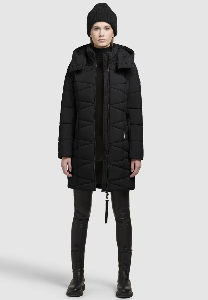 Zwarte pufferjas met een capuchon, mooi gewatteerd patroon en ritssluiting aan de voorkant. Gecombineerd met zwarte leggings en kniehoog laarzen. Minimalistisch ontwerp.