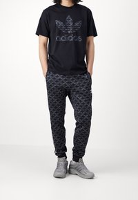 Svart t-shirt med Adidas-logotyp i bladmönster, paired med mönstrade svarta joggers och grå träningsskor.