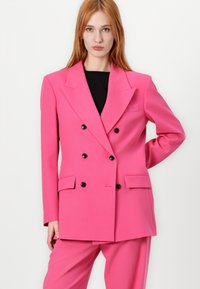 Roze dubbelzijdige blazer met een ingelegde kraag, zwarte knopen en zijzakken. Gecombineerd met bijpassende broek, beide in een gladde textuur.