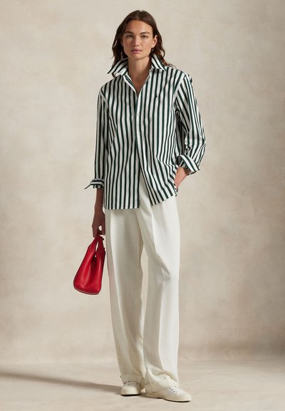 Frau trägt ein grün-weiß vertikal gestreiftes Hemd, weiße Hosen mit weitem Bein, weiße Turnschuhe, hält eine kleine rote Handtasche.