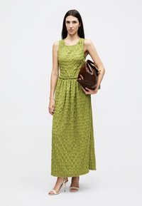 Γυναίκα που φοράει αμάνικο πράσινο μάξι φόρεμα με υφή, κρατώντας μεγάλο καφέ δερμάτινο φάκελο, φορεί λευκά τακούνια σανδάλια, στέκεται μπροστά σε απλό φόντο.