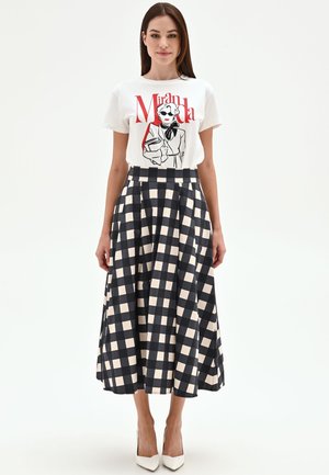 Femme debout portant un t-shirt blanc à motif avec texte et illustration rouges, associé à une jupe midi à carreaux noirs et blancs taille haute et des talons blancs.