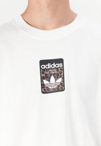 White cotton T-shirt featuring a black rectangular logo with leopard print design and the Adidas trefoil emblem. Classic crew neck style.
