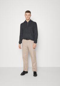 Mørk grå ribbestrikket genser med halv-zip krage, kombinert med lyse beige bukser og svarte sko med rynkete detaljer.