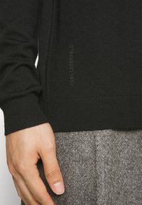 Gros plan sur une main reposant près de l'ourlet d'un pull noir en maille avec le logo "KARL LAGERFELD", associé à un pantalon gris texturé.